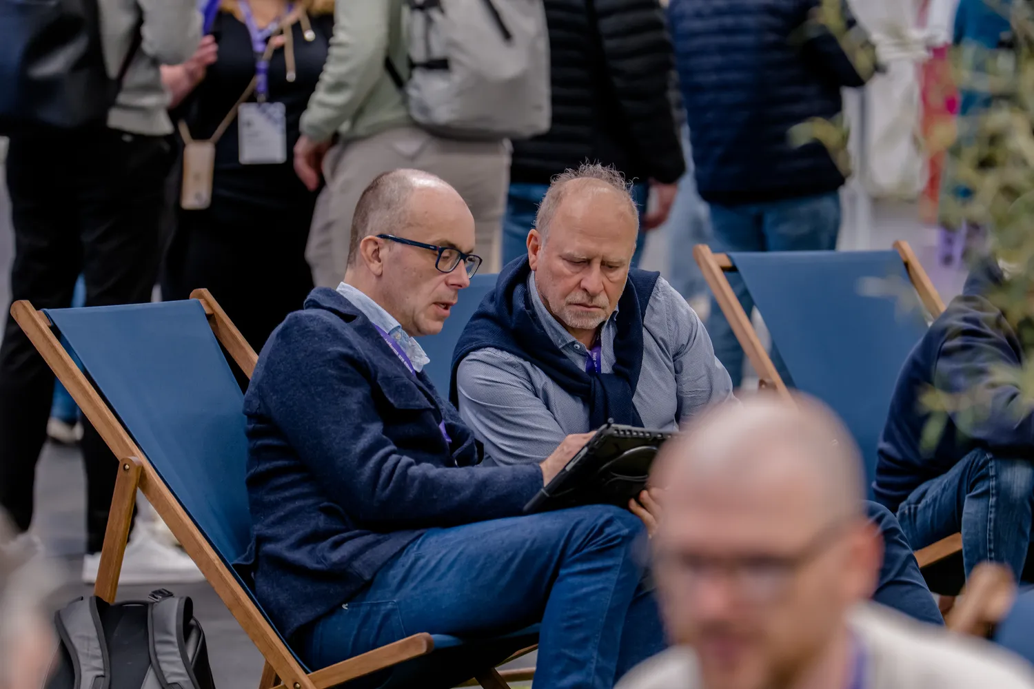 Two men seated on folding chairs at an outdoor conference event, taking notes while surrounded by other attendees. They are dressed in business casual attire, and the background shows blurred participants engaged in discussions.