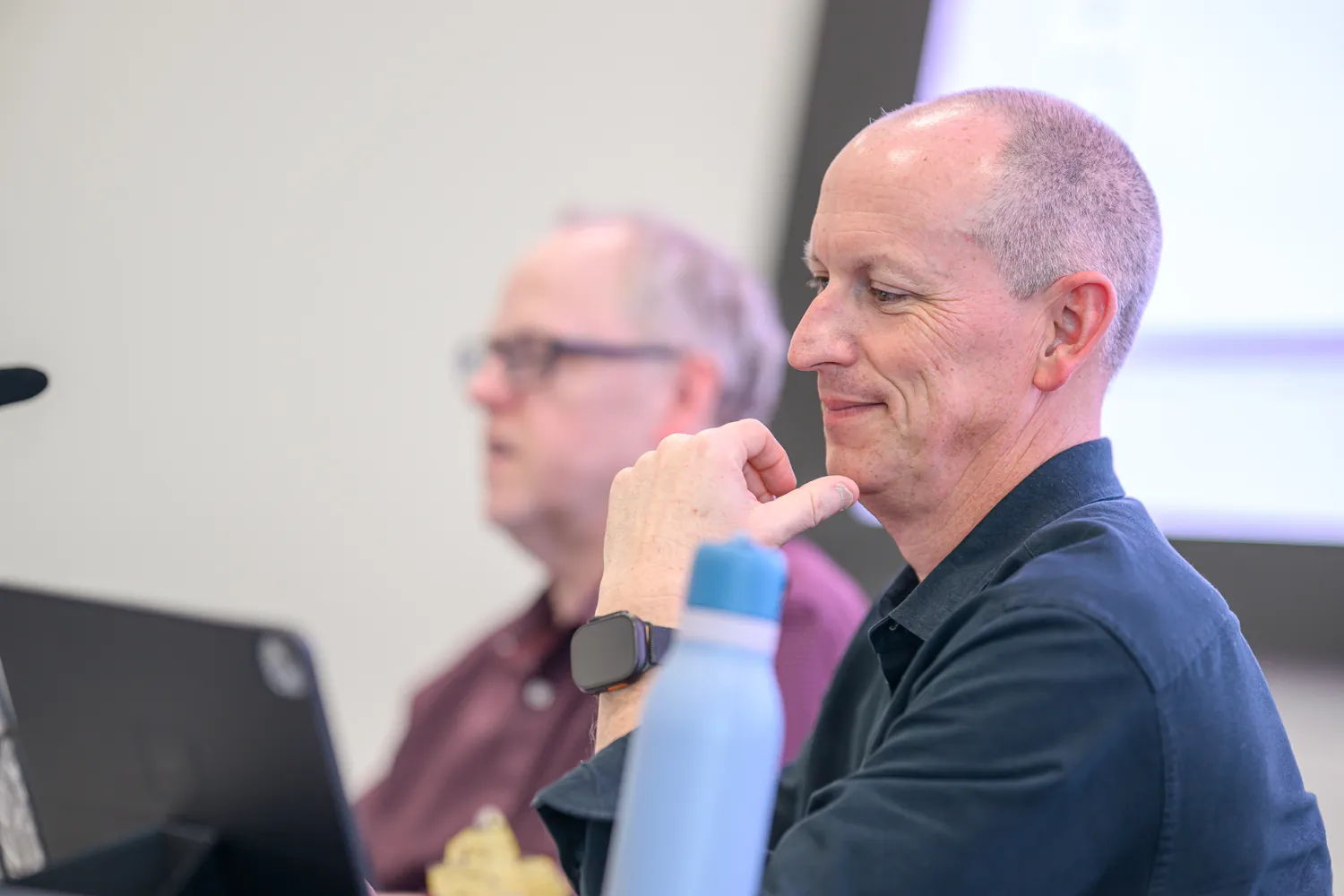 A man in a dark shirt gestures while speaking to an audience, engaging attentively. The background shows another person in a blurred focus, seated and listening.