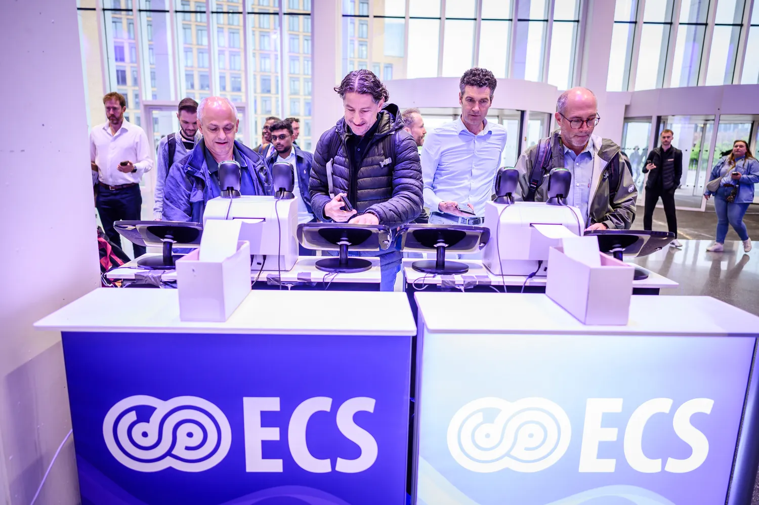 Attendees gather at a branded booth displaying the logo of COECS, engaging in signing activities with promotional items positioned on a sleek display counter in an indoor conference venue. The scene captures professional individuals interacting near modern, well-lit exhibition space.