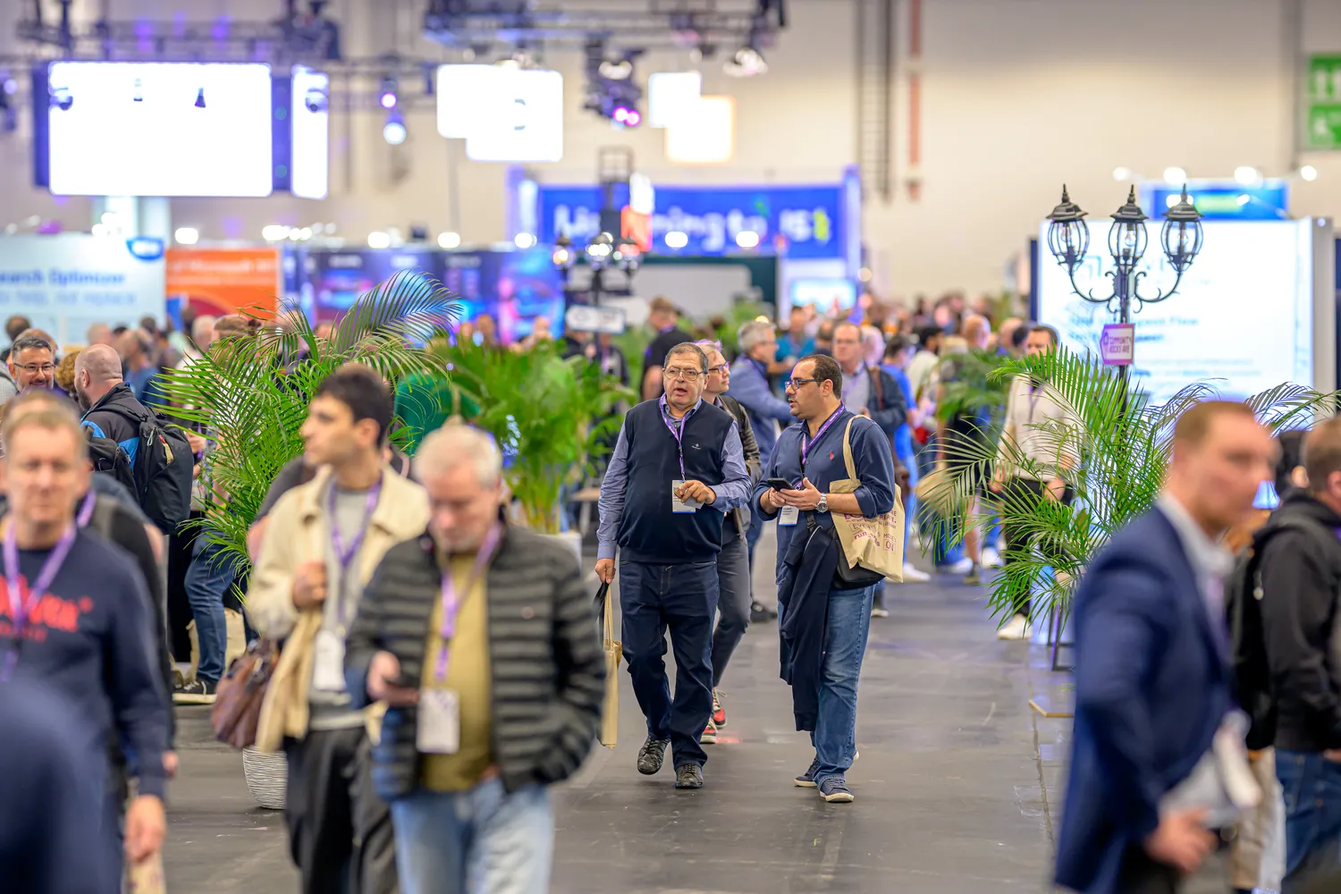 Attendees at a professional conference walk through a well-lit indoor corridor lined with decorative plants and modern lighting fixtures. The scene captures diverse participants engaged in discussions, surrounded by various exhibition booths and digital displays.