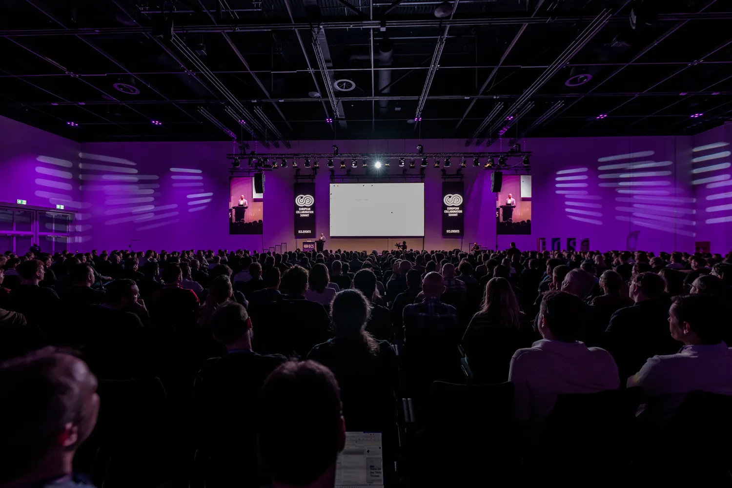 A large audience attentively faces a stage with a speaker delivering a presentation. The stage features prominent branding elements, including illuminated logos and dynamic lighting effects.