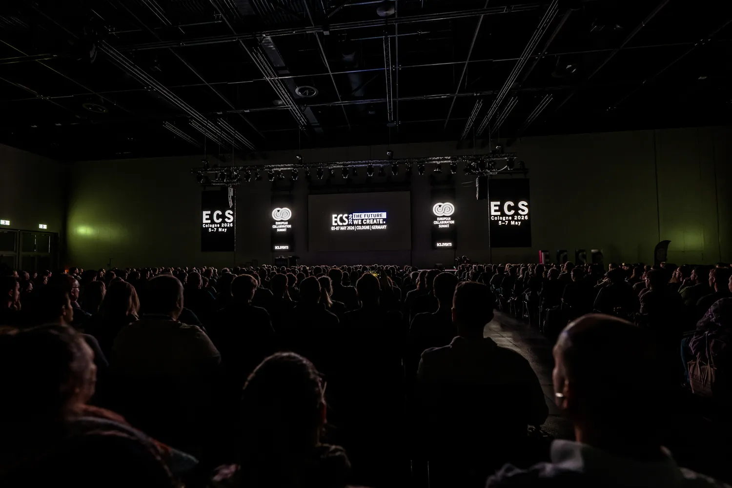 A large audience seated facing a stage at the ECS Cologne 2026 event, displaying branding and event details including dates and location. The backdrop features prominent screens with logos and promotional text about innovation in the creative industry.