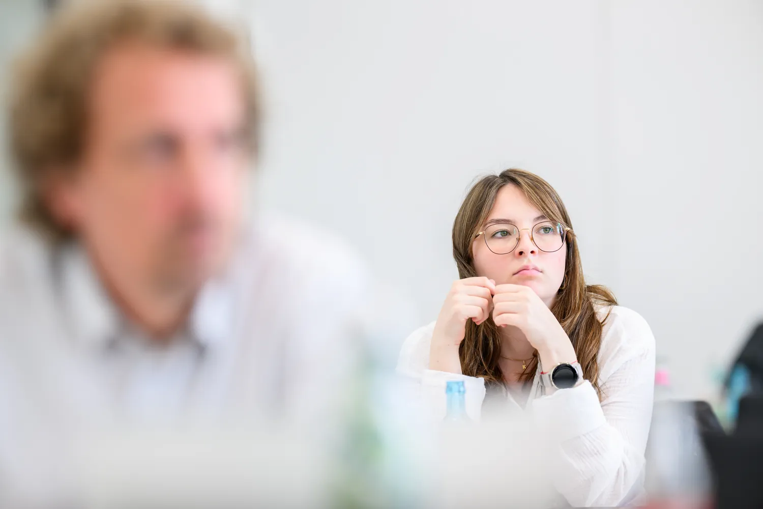 A woman in professional attire with glasses holds a marker, appearing to engage in a discussion or presentation. She is positioned near another individual, suggesting a collaborative or seminar setting.