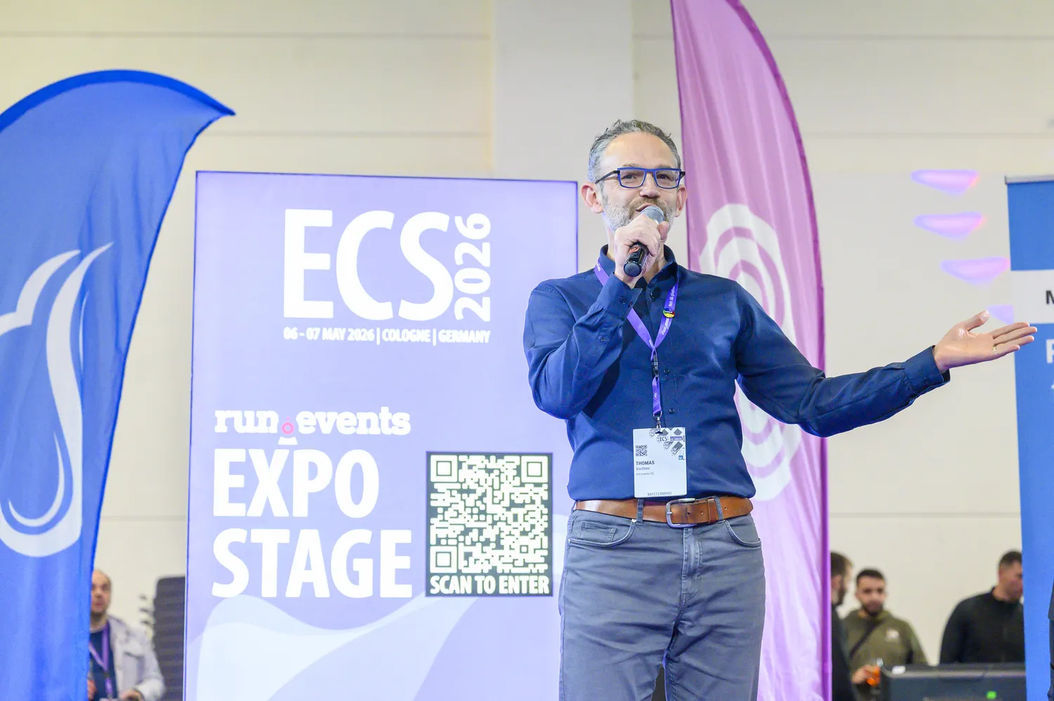 Man speaking at an event stage during ECS 2026, holding a microphone and gesturing with his hands. The backdrop displays the event name, dates, location in Cologne Germany, and a QR code for entry.