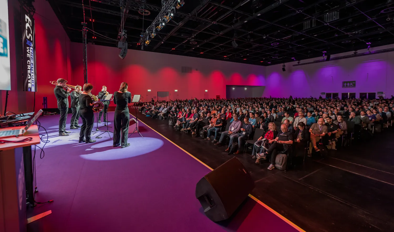 A large audience attends an indoor event featuring an orchestral performance on stage with brass instruments and sheet music. The venue is brightly lit with vibrant stage lighting, including red and purple hues.