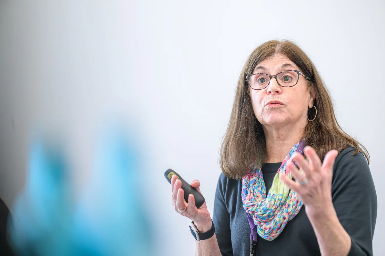 A woman in professional attire with glasses and a colorful scarf speaks to an audience, gesturing with her right hand while holding a microphone or handheld device. She appears engaged and delivering an informative presentation in what looks like a conference or seminar setting.