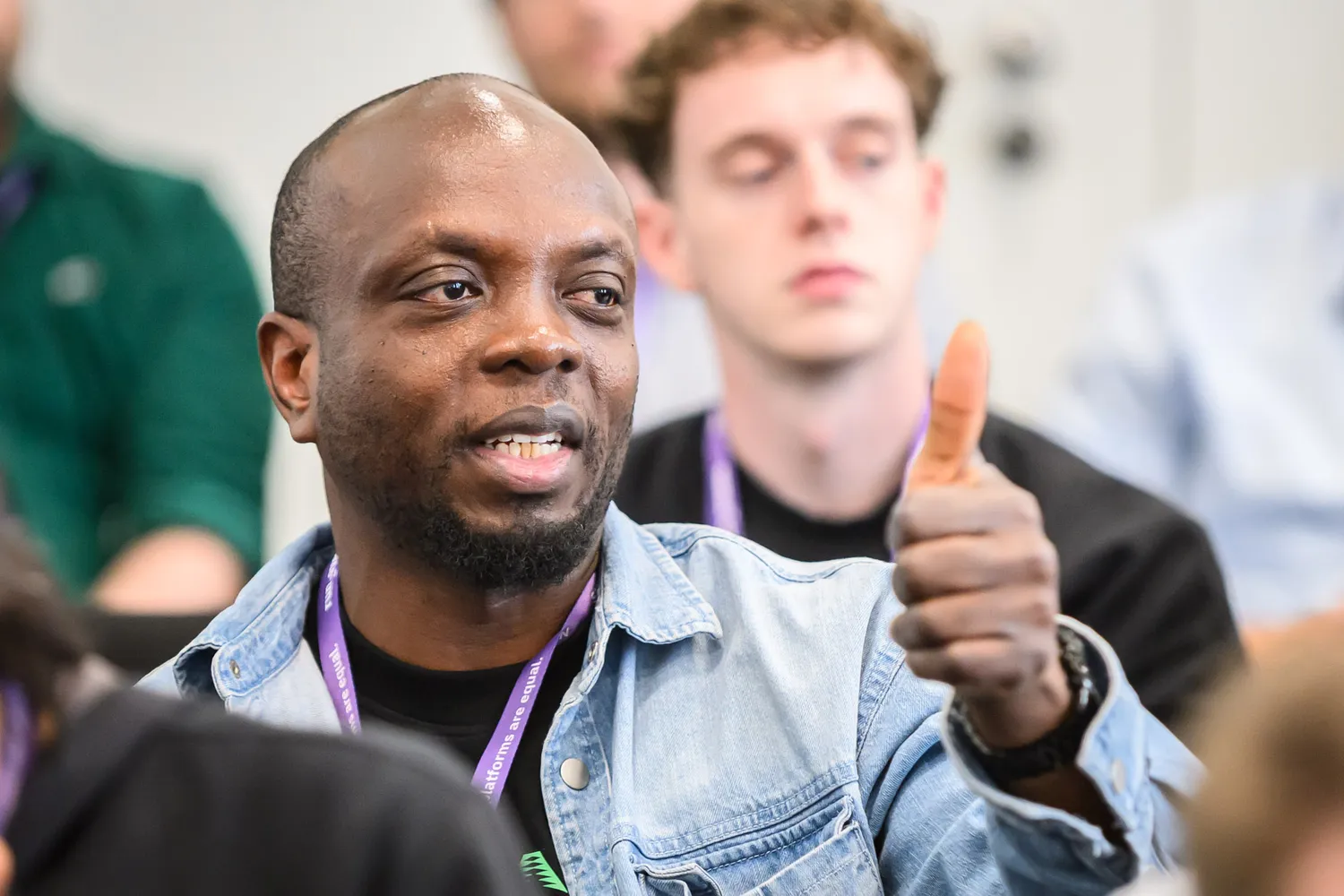 A man in a denim jacket and lanyard gestures positively with his thumb up, engaging an audience at what appears to be a professional conference or seminar. The background shows blurred attendees, indicating an interactive presentation setting.