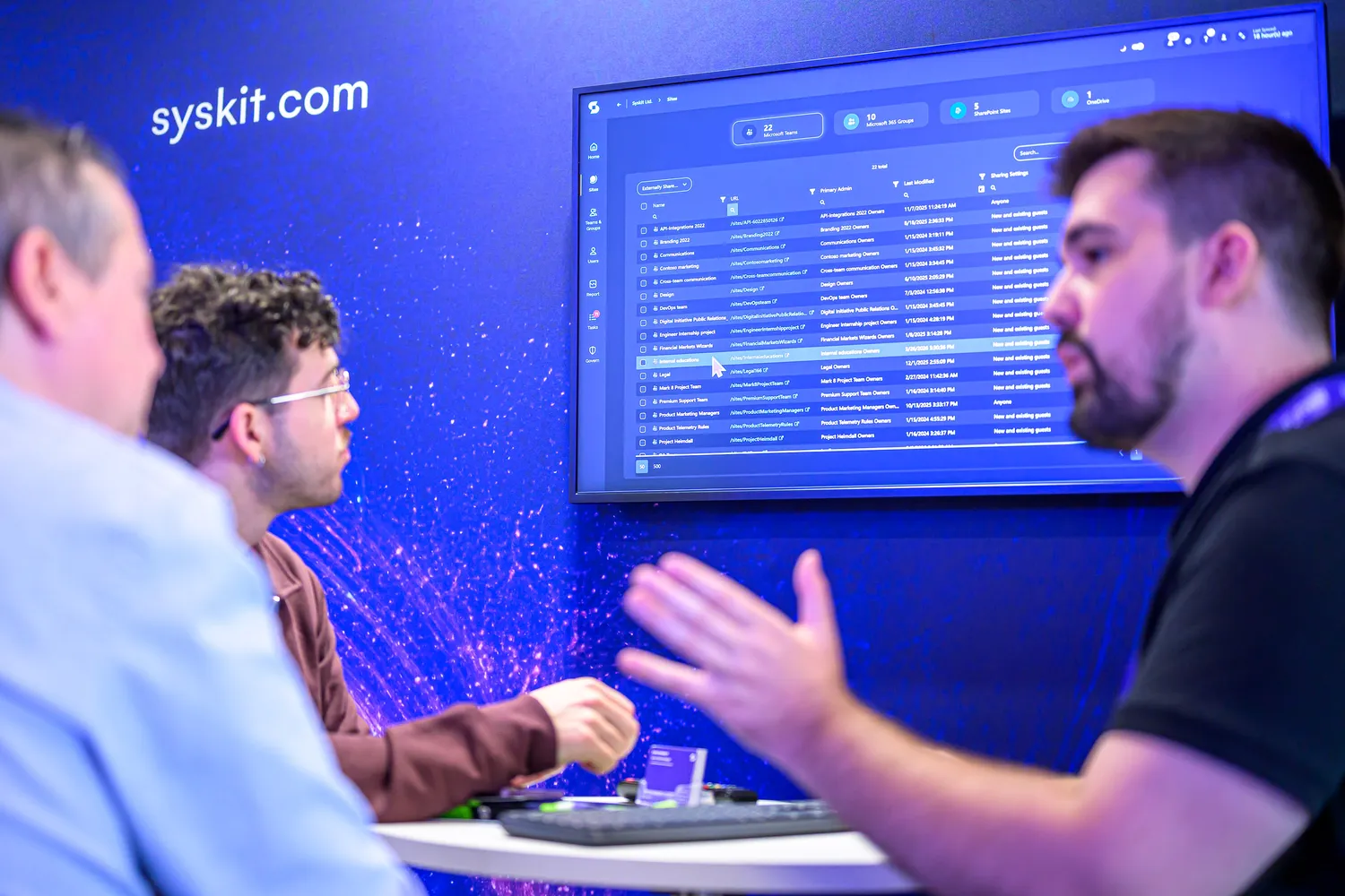 Professional team members discuss project management tasks on a digital interface displaying Syskit’s team organization and task lists. The scene highlights collaborative workflows and project coordination within a tech-focused environment.