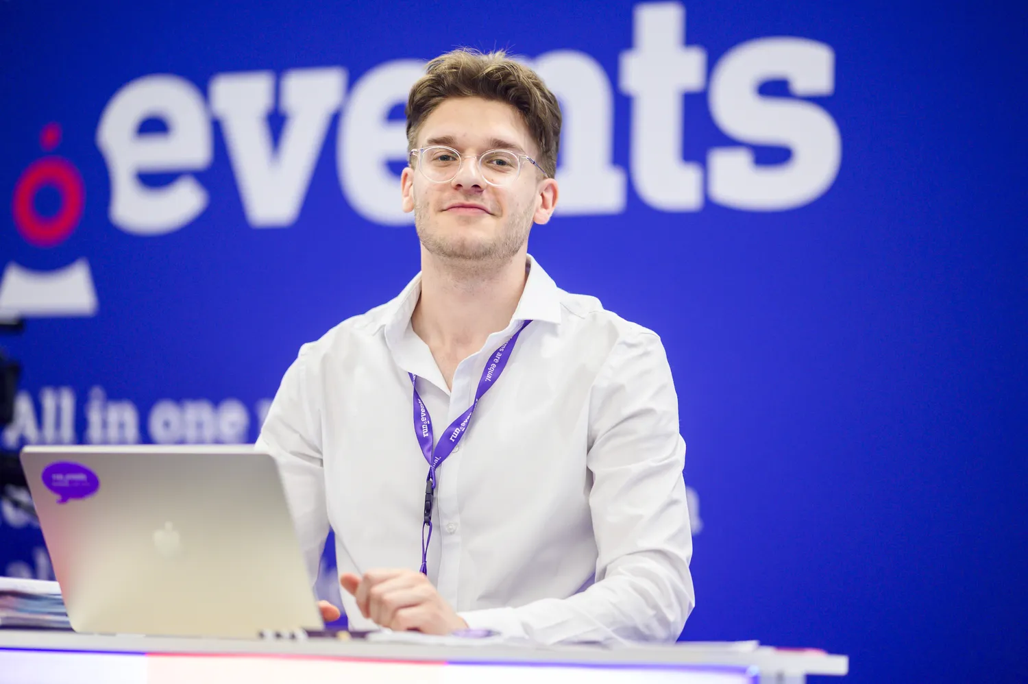 A person in a white shirt and lanyard presents or interacts with a laptop at an event booth. The backdrop features bold text and branding elements related to event management.