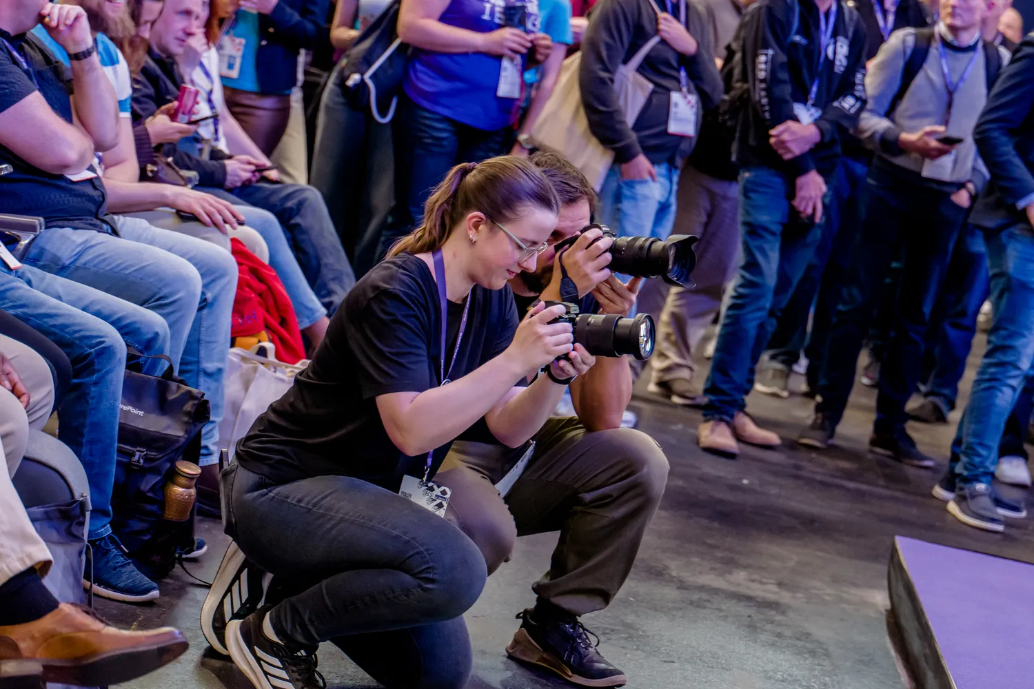 A photographer kneels and focuses on capturing images of engaged attendees at a professional event. The scene includes individuals wearing lanyards, casual business attire, and some holding devices like phones.