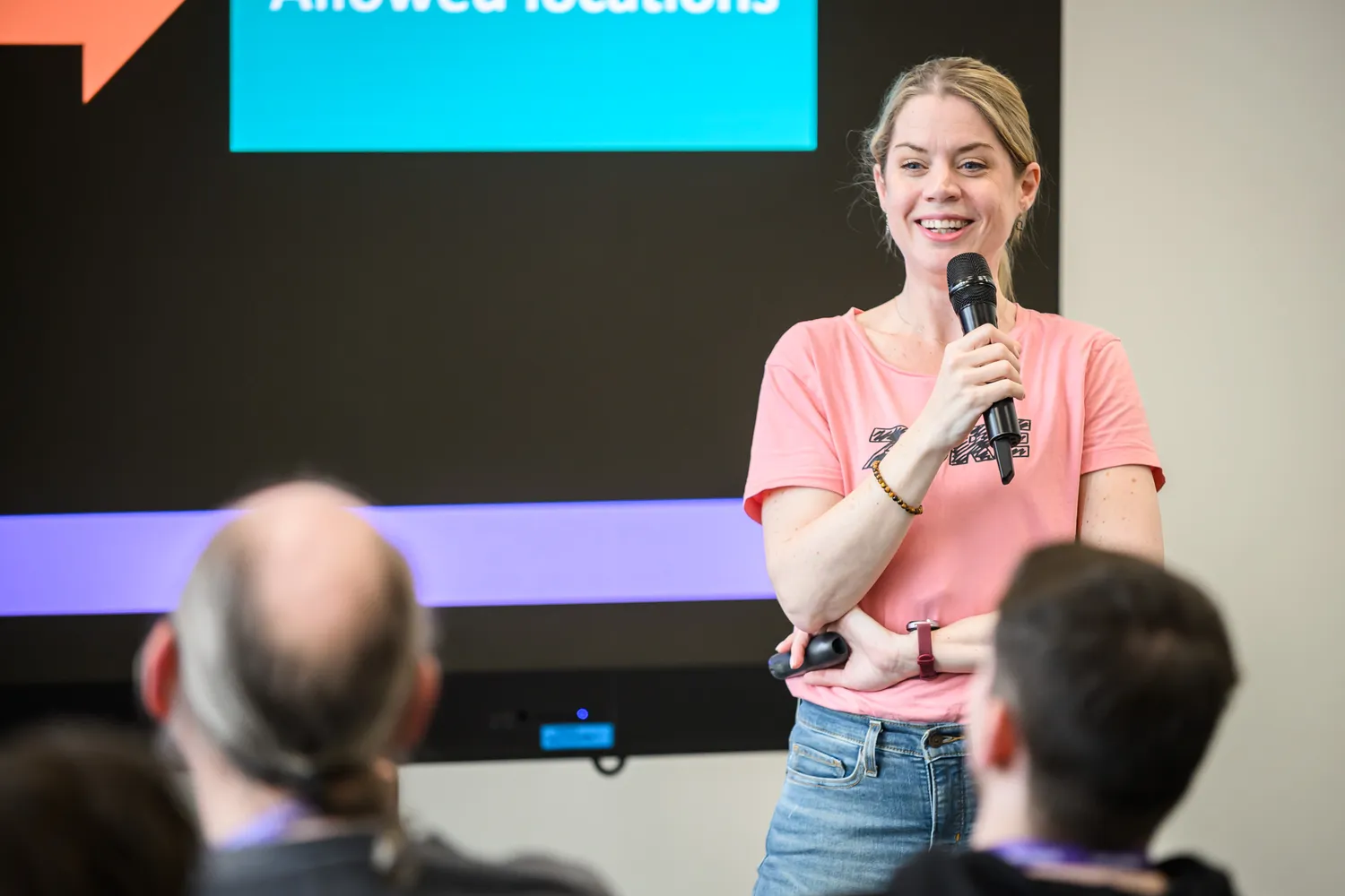 A woman in a casual pink shirt speaks into a microphone at an indoor event, engaging an attentive audience. The background includes a modern screen displaying partial text and vibrant geometric lighting.