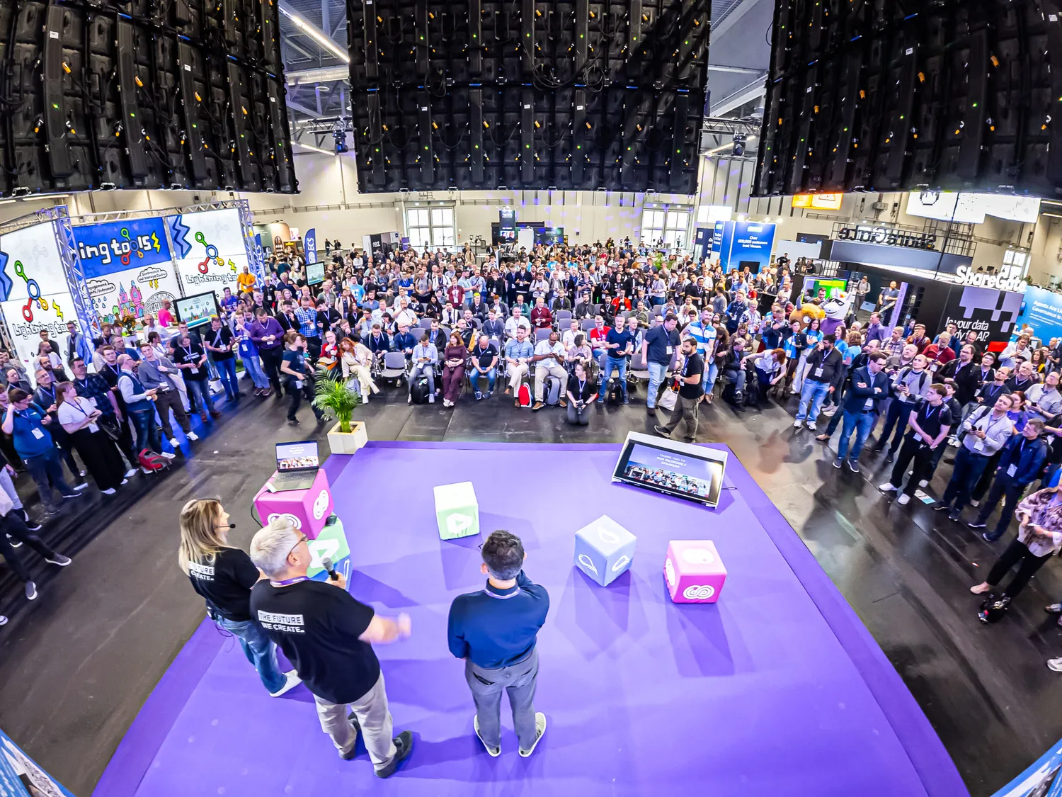 A bustling tech expo venue features an engaged audience gathered around a vibrant interactive demo station. Presenters and participants interact with a digital setup, including cubes and screens, while colorful booths and event lighting enhance the lively atmosphere.