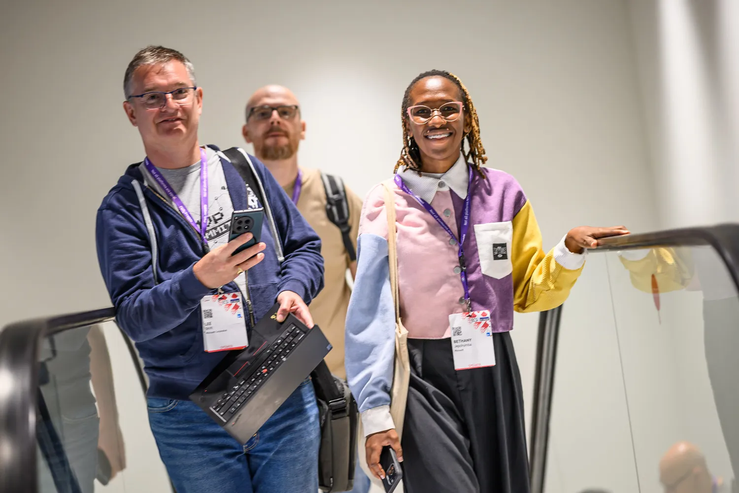 Three individuals at a professional conference or trade show, smiling and engaged. One person holds a laptop, while another wears a blazer and name badge, indicating participation in networking or product demonstrations.