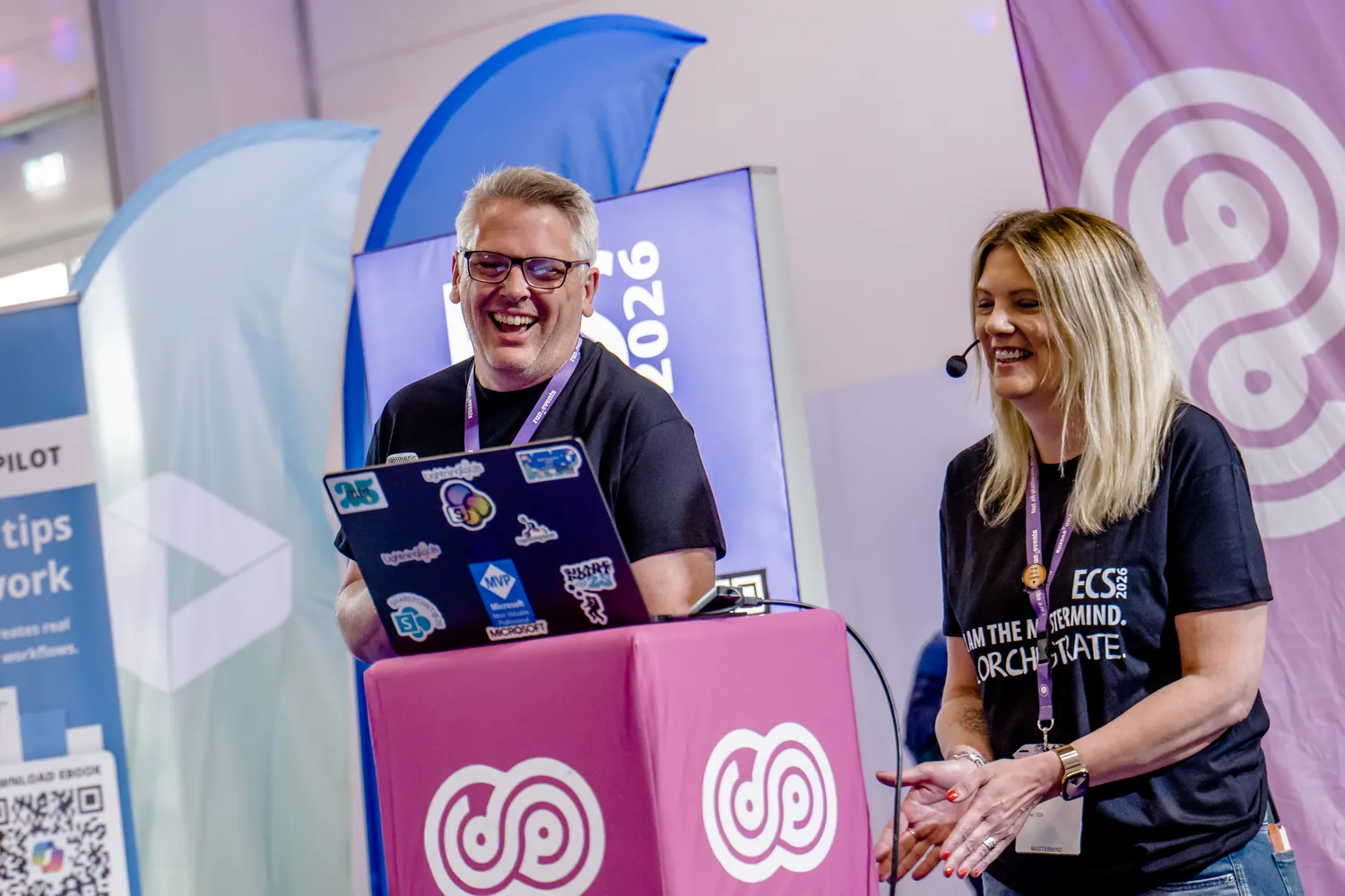 Two individuals present at a tech conference booth, holding a decorated laptop and engaging in a discussion. The backdrop features event branding and promotional materials for digital workflow solutions.