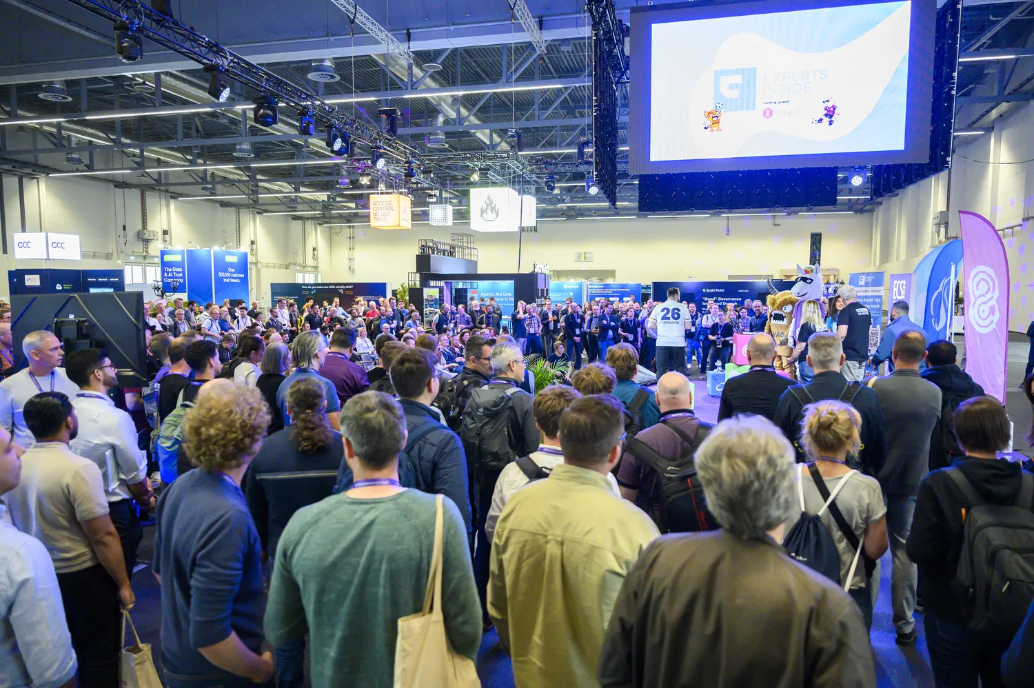 A bustling professional conference hall features numerous attendees facing forward, engaged in a presentation or panel discussion. The scene includes branded banners, stage lighting, and participants dressed in business and casual attire.