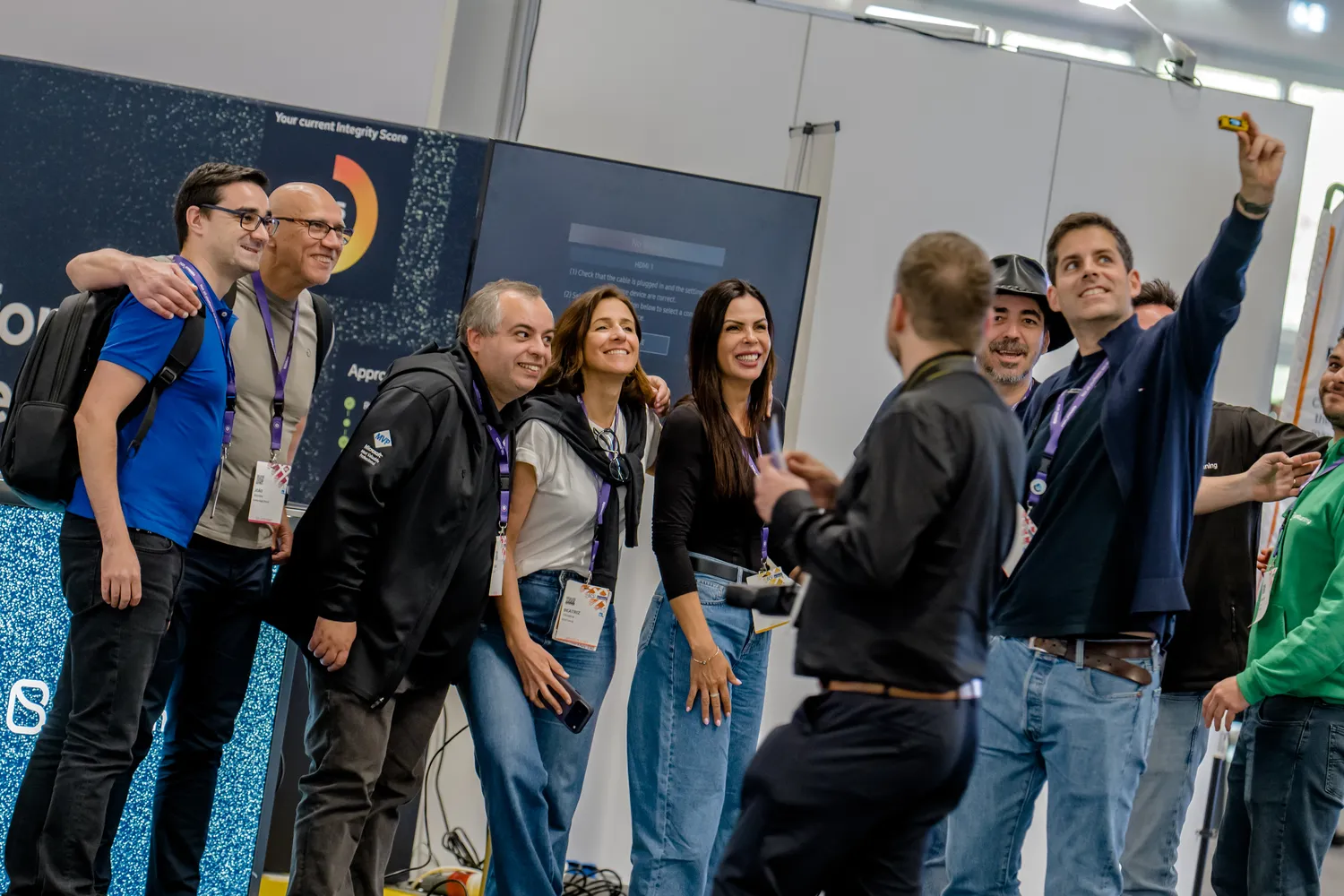 Attendees at a professional conference or expo gathered around a digital display, smiling and interacting enthusiastically. The scene includes badges, casual business attire, and an interactive setup with text prompts on the screen.