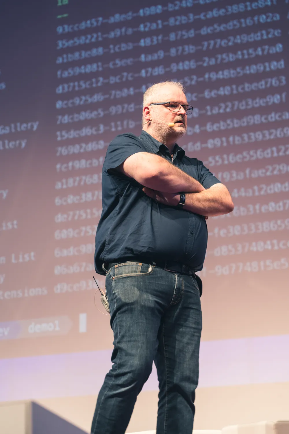 Man in business attire stands confidently with arms crossed, presenting at a tech conference. Digital code and data patterns form a vibrant backdrop behind him.