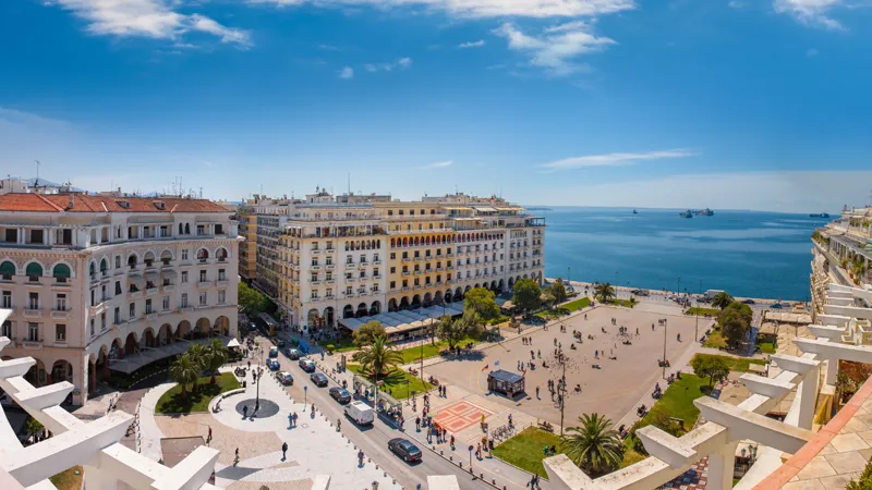 A vibrant coastal urban plaza featuring historic multi-story buildings with classical architectural details. The scene includes a spacious open area with people walking, a waterfront view of the sea, and lush greenery along pathways.