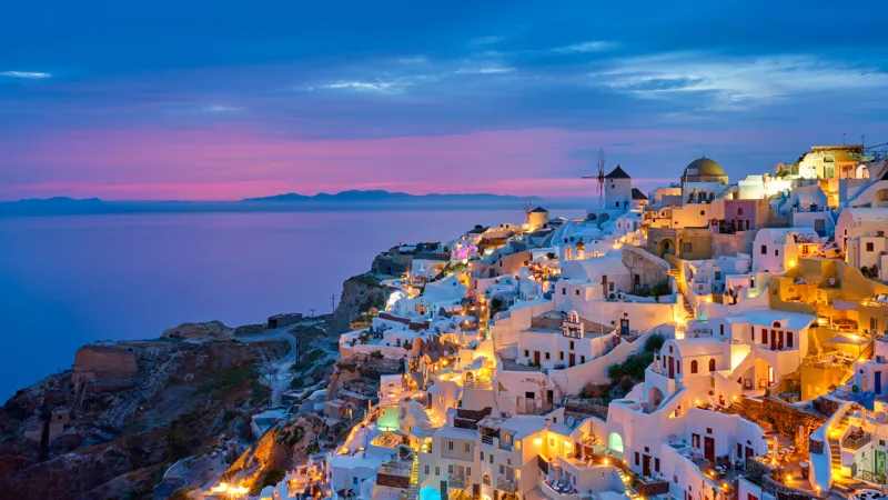 A coastal village with white-washed buildings and domed roofs on cliffs at twilight, with warm lights glowing above a calm sea.