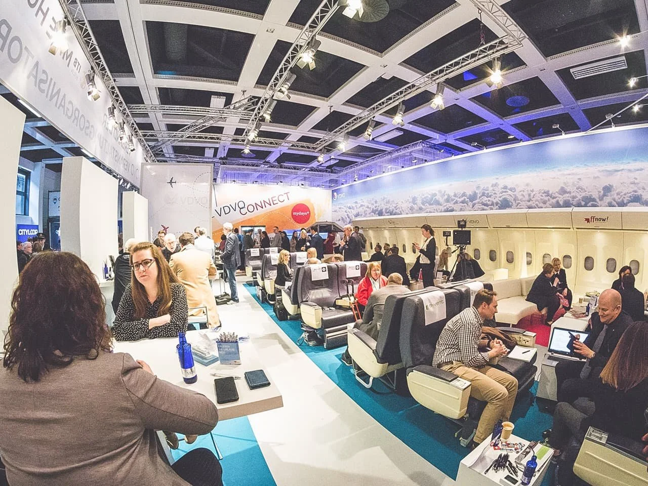Professional attendees gather at a trade show booth designed to resemble an airplane cabin. The setup includes seating arrangements, laptops, and promotional displays featuring aviation and connectivity branding.
