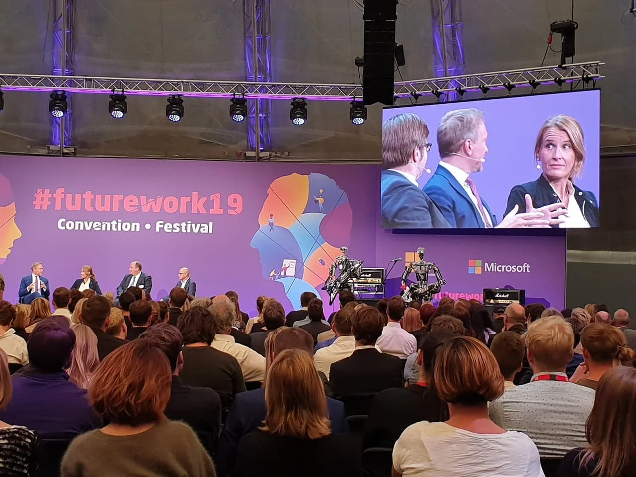 A professional panel discussion at the FutureWork19 convention featuring four speakers engaged in conversation. Attendees are seated attentively in front of a vibrant stage backdrop with colorful abstract silhouettes and branding elements, including a live screen displaying the panelists.