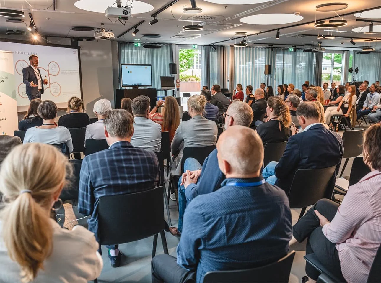 Attendees seated in a modern conference room listening to a speaker at the front, engaging in what appears to be an educational or business presentation. The room features professional lighting, presentation screens, and participants dressed in business attire.
