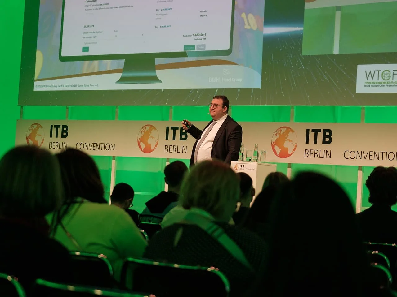 A professional speaker at ITB Berlin Convention delivers a presentation to an audience, with a digital pricing display projected behind them. The event features branding from ITB and WTOF, and attendees are seated attentively.
