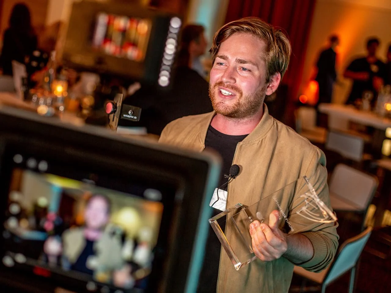 A man in a brown jacket presents an award trophy at what appears to be a lively evening event. The background shows blurred attendees and decorative lighting, suggesting an engaging social or networking atmosphere.