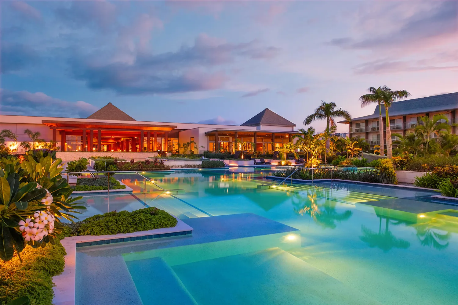 Elegant resort setting featuring a large, illuminated infinity-style pool surrounded by lush greenery and tropical plants during twilight. Warmly lit buildings with architectural details and palm trees create a sophisticated, inviting atmosphere.