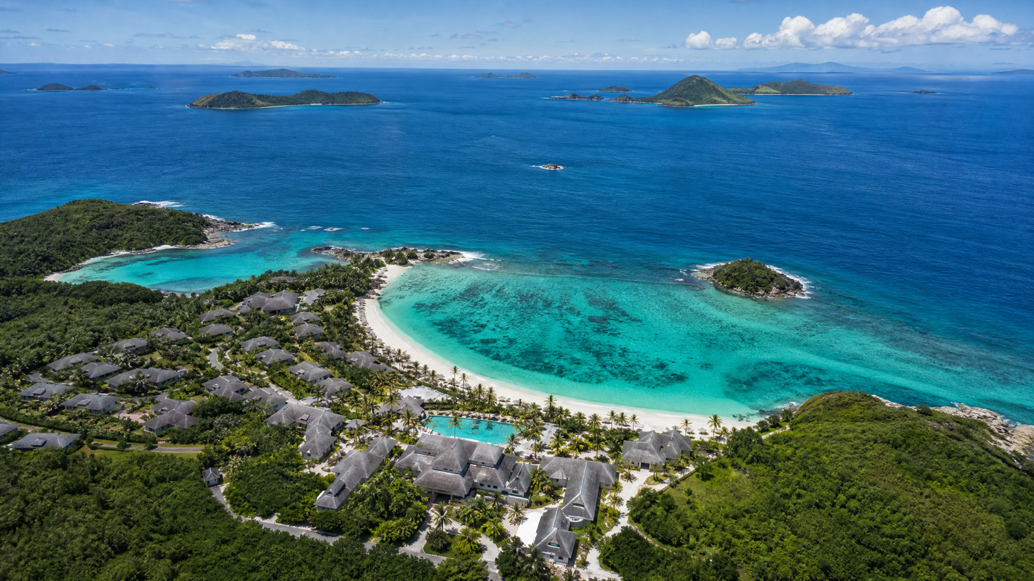 A serene aerial view of a tropical resort surrounded by lush greenery, featuring traditional-style buildings and a private, curved beach with stunning turquoise waters. The resort is nestled near distant islands under a clear sky.