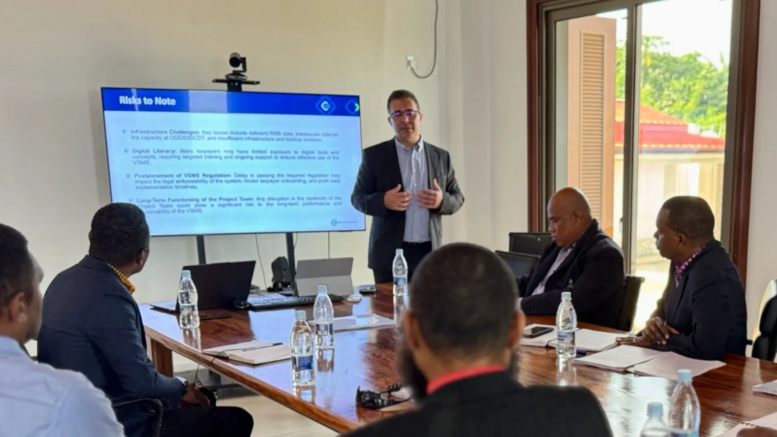 A professional presentation session where a speaker discusses key risks related to project challenges. Attendees are seated around a table, engaged in the discussion, with water bottles and laptops visible.
