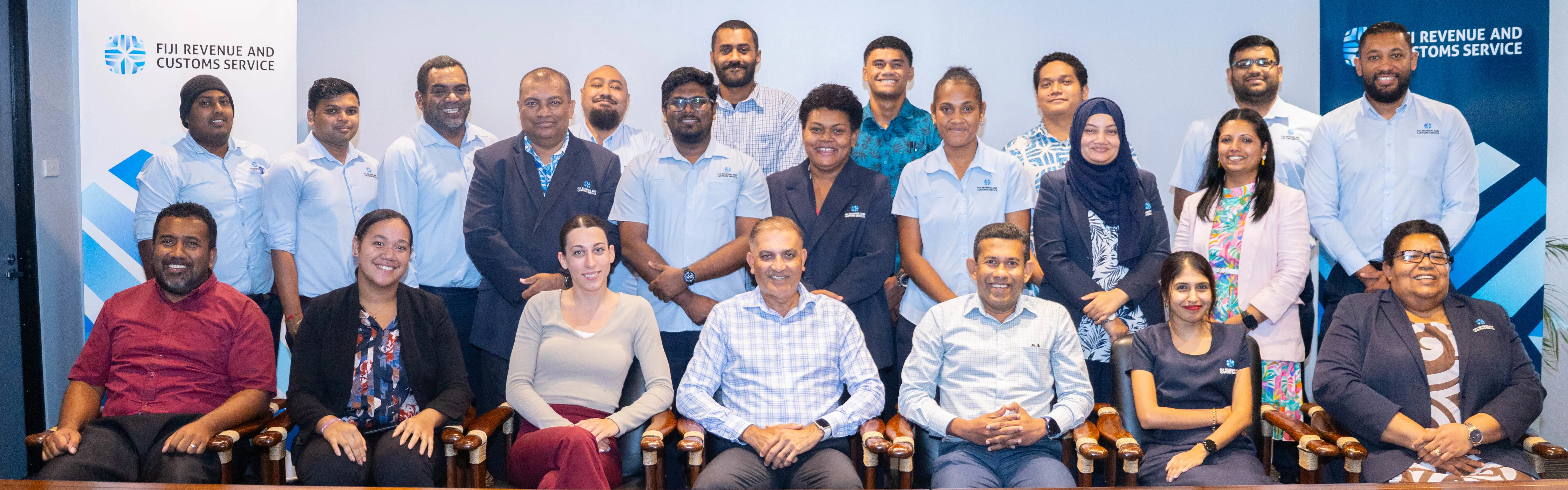 Professional team members of Fiji Revenue and Customs Service posing together in a formal setting, seated and standing behind a backdrop with the organization’s logo. The group includes individuals in business attire, reflecting unity and collaboration.