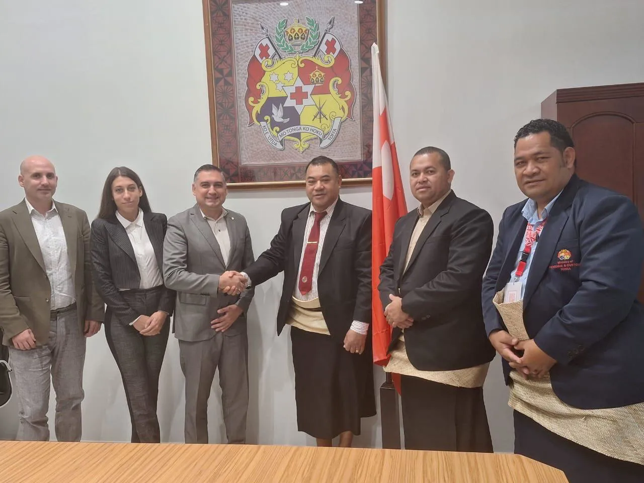 Six individuals in formal attire pose for a professional photo, with one central figure shaking hands. The backdrop features an official coat of arms and a national flag, indicating an official or diplomatic setting.