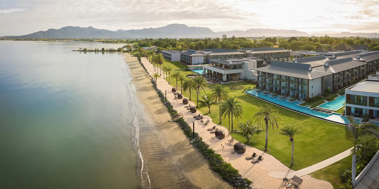 Elegant resort complex featuring expansive lagoon views, modern buildings, and lush tropical landscaping along a sandy beach path with outdoor seating and palm trees. Outdoor swimming pools and scenic mountain backdrops enhance the tranquil and luxurious setting.