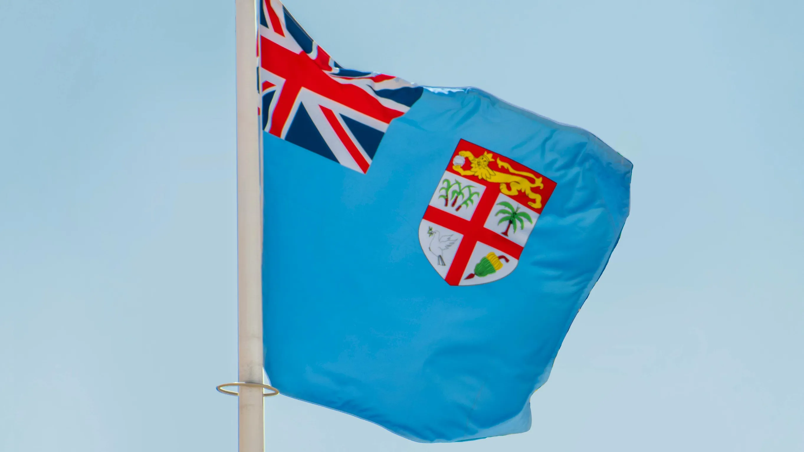 A blue flag with the Union Jack at the upper hoist side and a coat of arms featuring palm trees, a shield, and other symbols is prominently displayed against a clear sky. The flag appears to be waving gently on a flagpole.
