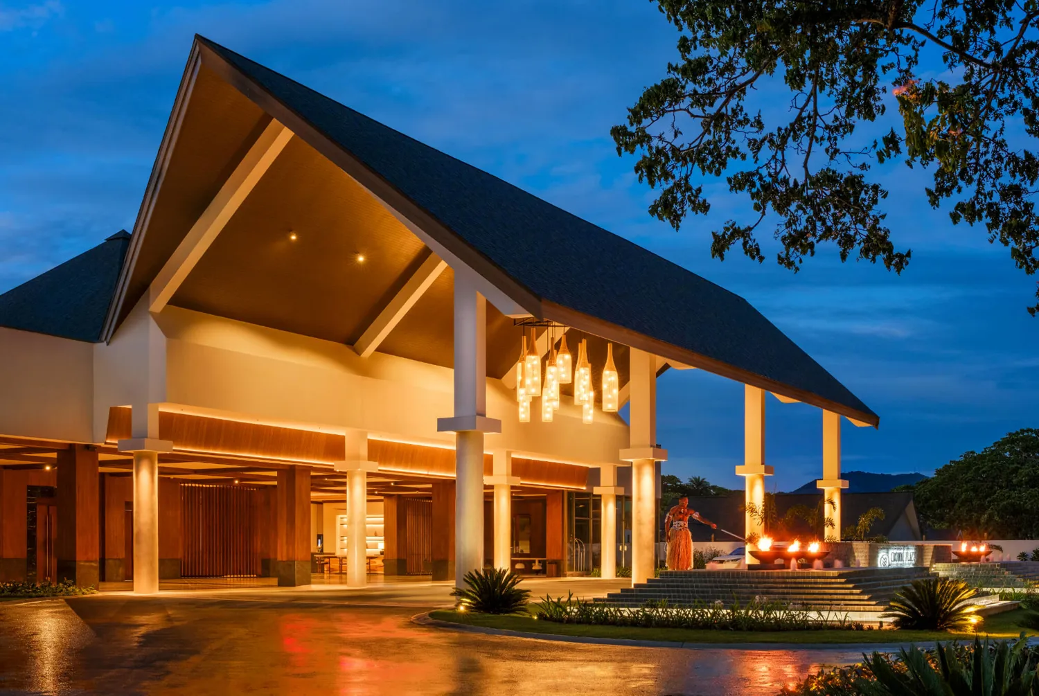 Modern architectural building entrance illuminated at dusk with warm lighting, featuring large pillars, decorative hanging lights, and a person standing near a fire pit in the foreground. The scene exudes a sophisticated, inviting atmosphere with lush greenery and a serene evening sky.