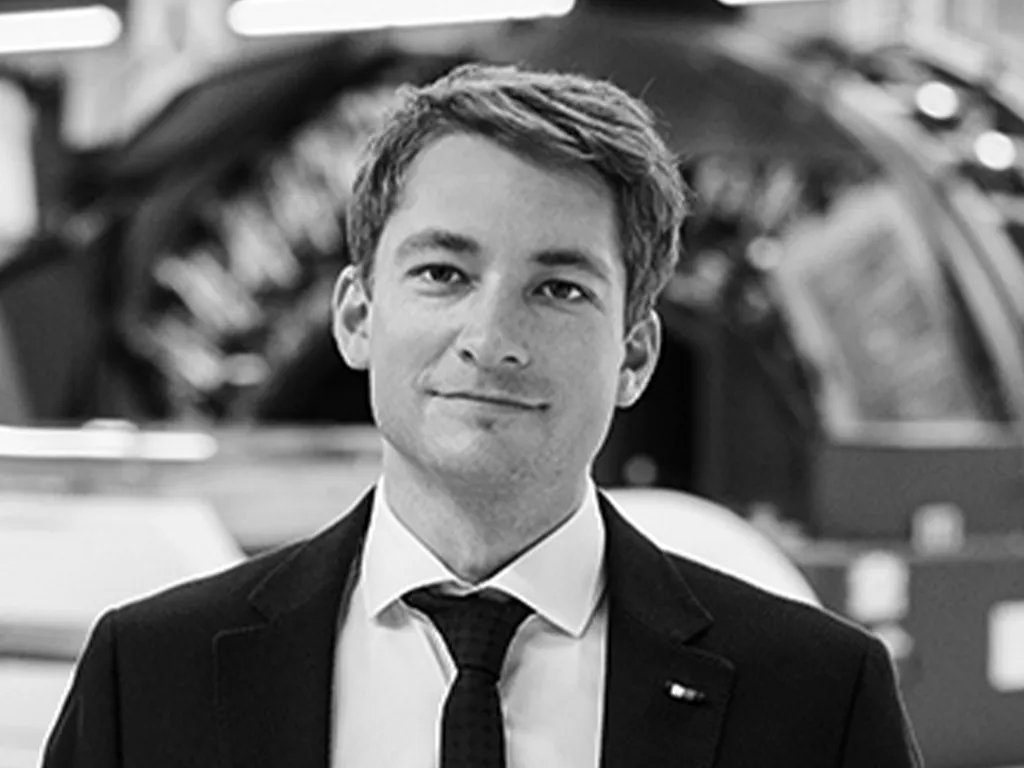 A man in formal attire, wearing a suit jacket, dress shirt, and tie with a small emblem on the lapel, poses professionally. The background suggests an indoor setting with blurred industrial or office elements.