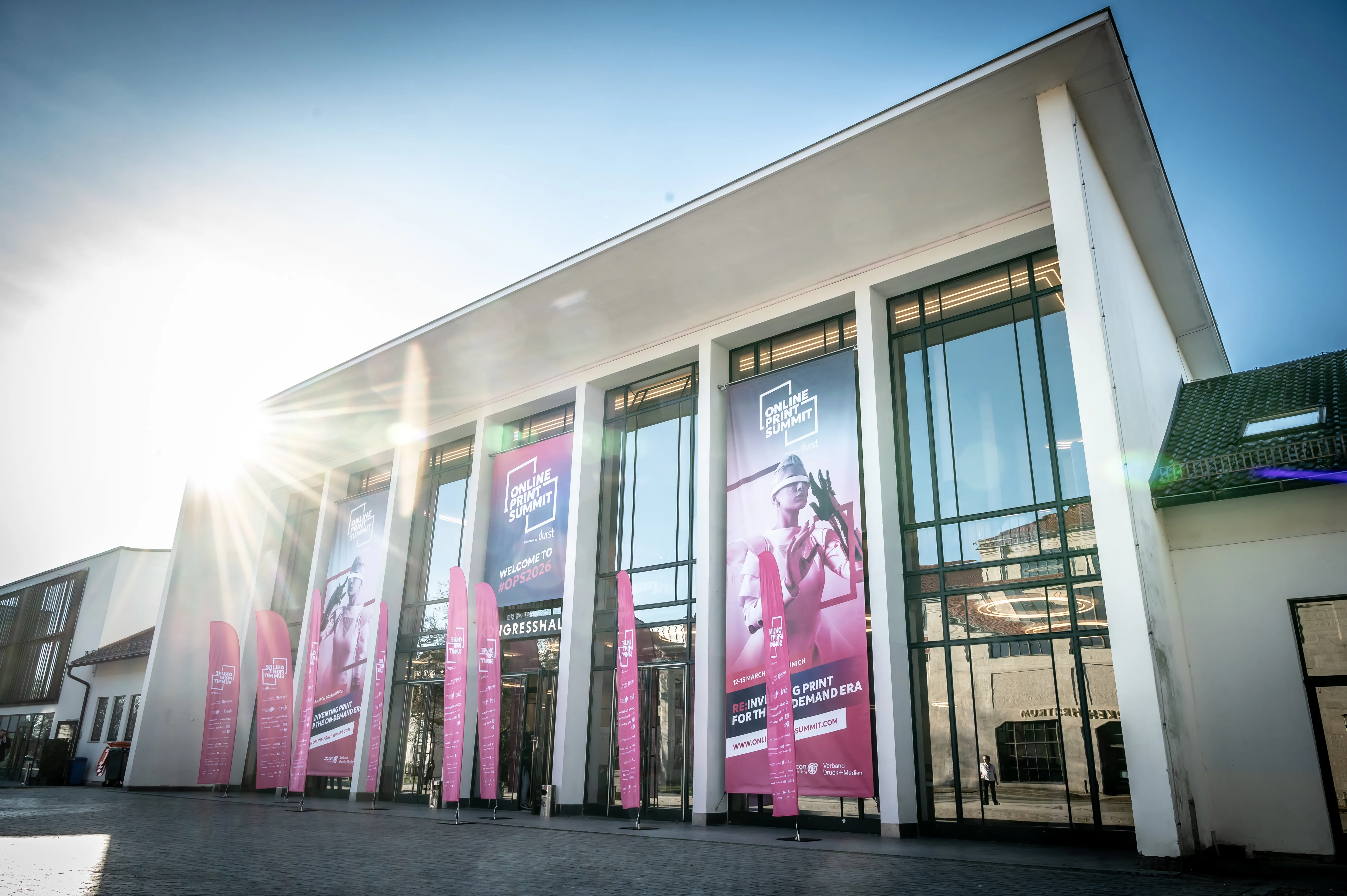 Modern conference building exterior with large glass entrance doors displaying promotional banners for the Online Print Summit