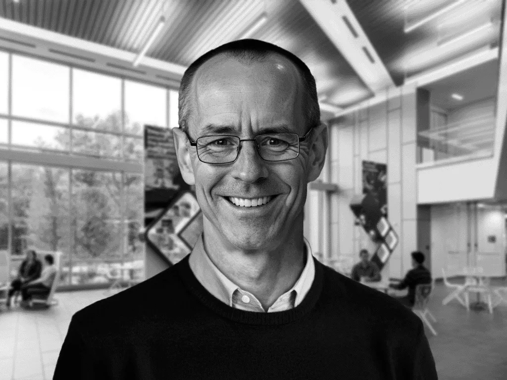 A man wearing glasses and a dark sweater smiles warmly in an indoor conference or meeting space. The background features modern office elements, including glass walls, abstract wall art, and a few people seated in the distance.