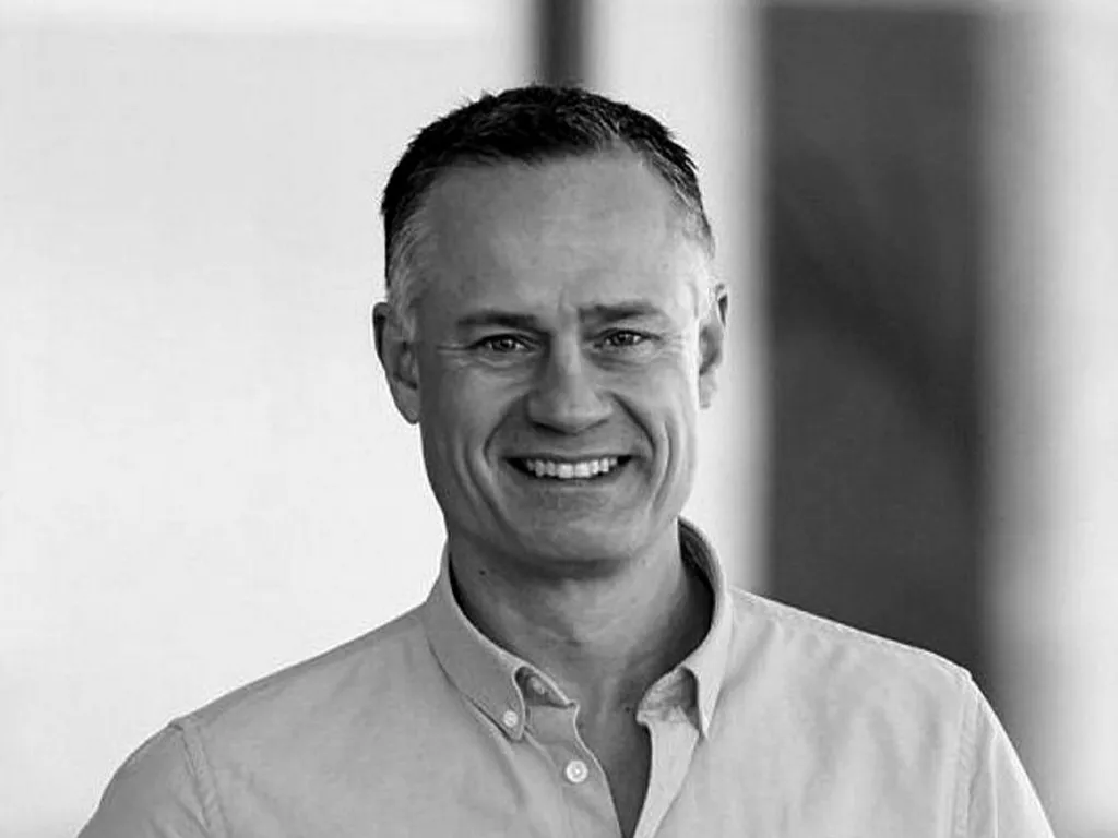 A man in a light-colored button-down shirt smiles warmly at the camera against a neutral background. He appears in a professional setting, exuding approachable confidence.