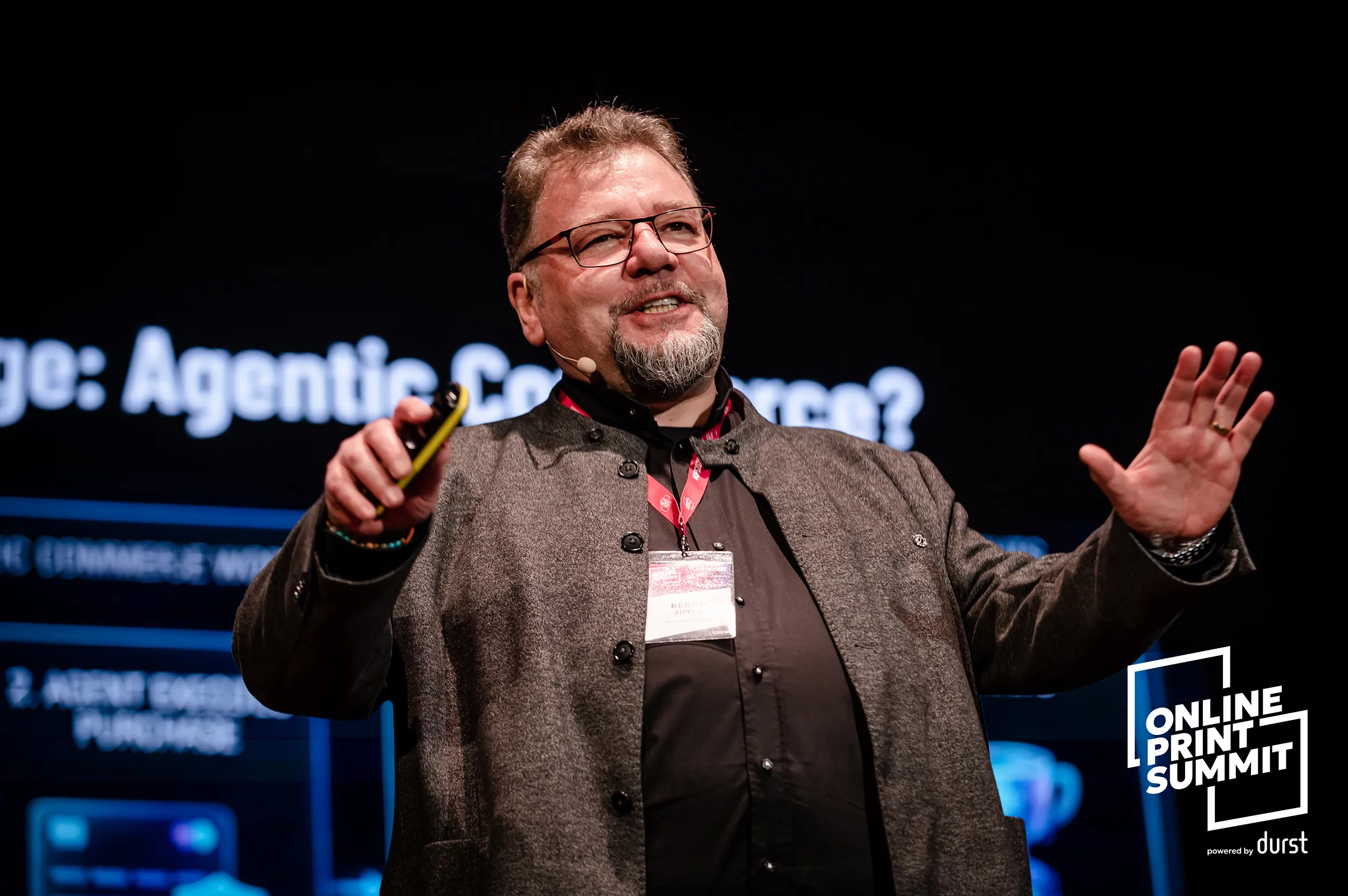 A speaker presents on stage at the Online Print Summit, gesturing toward a screen during a conference session.