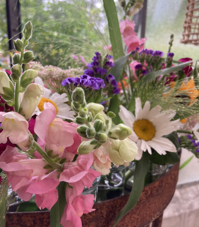 A decorative floral arrangement featuring vibrant pink snapdragons, white daisies, purple flowers, and lush greenery arranged in a rustic container. The bouquet includes natural foliage and subtle outdoor lighting elements.