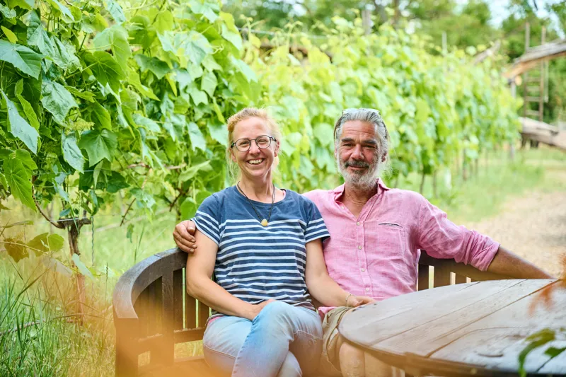 Two individuals seated outdoors on a wooden bench surrounded by lush vineyard greenery, smiling warmly at the camera. They are dressed casually in striped and checkered shirts, enjoying a serene rural setting.