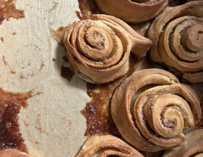 Close-up of golden-brown cinnamon rolls with swirled filling, placed on a light-colored parchment. The pastries exhibit a slightly caramelized glaze and appear freshly baked.