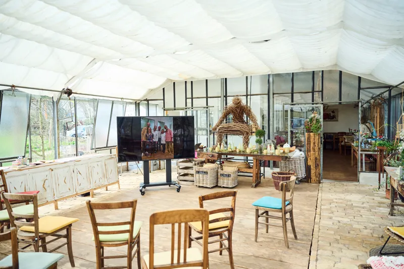 Indoor event venue featuring a spacious, well-lit area with wooden seating arrangements and rustic decor. A display screen shows a group photo, while decorative wicker baskets and natural elements like woven arches enhance the ambiance.