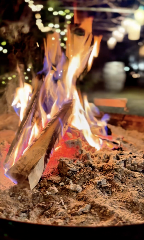 A close-up of burning logs emitting vibrant flames and embers against a dark background, highlighting the dynamic interplay of light and fire. The scene captures the warmth and intensity of a fire pit or bonfire.