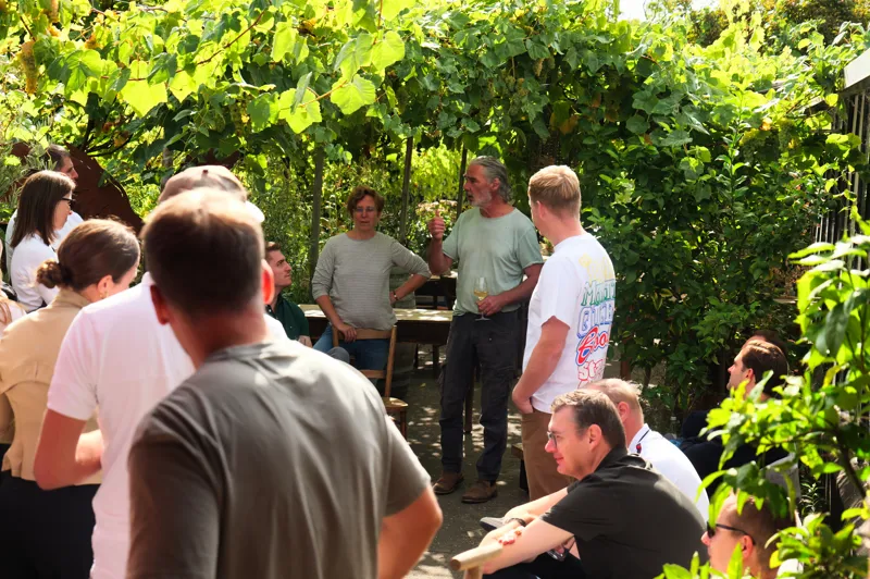 A group of people engaged in an outdoor discussion and tasting session surrounded by lush greenery. Participants are seated and standing around tables, with one individual holding a glass of what appears to be wine.