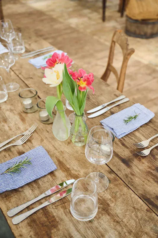 A rustic wooden table setting features neatly arranged cutlery, glasses, and blue-striped napkins. Vibrant floral arrangements in clear vases add a touch of natural elegance to the outdoor event setup.