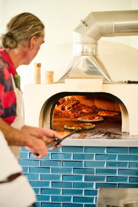A chef handles freshly baked pizzas in a high-temperature wood-fired oven, surrounded by a tiled kitchen backdrop. The scene captures the art of traditional pizza making with glowing flames and freshly cooked pizza slices.