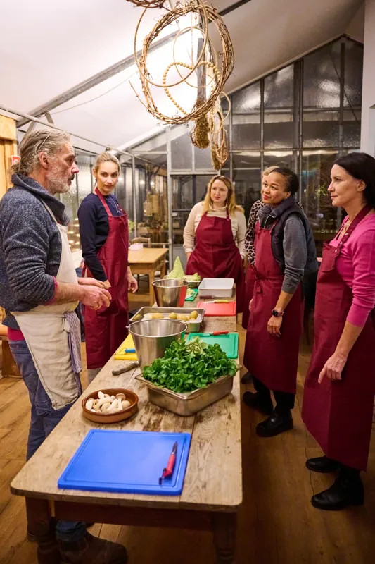 Group preparing food together in a rustic kitchen with herbs and ingredients on the table
